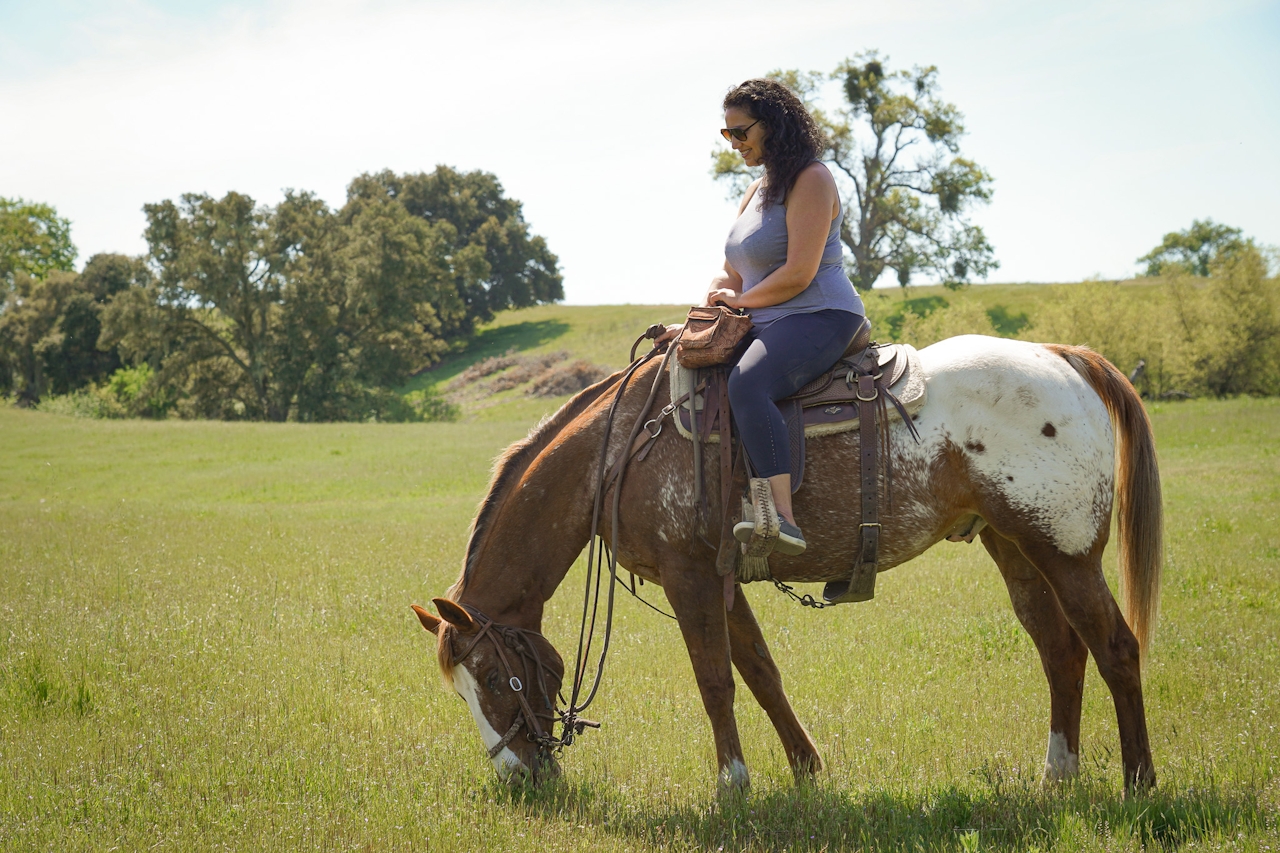 A woman on a horse in Santa Margarita Ranch sits and waits while her horse snacks on grass